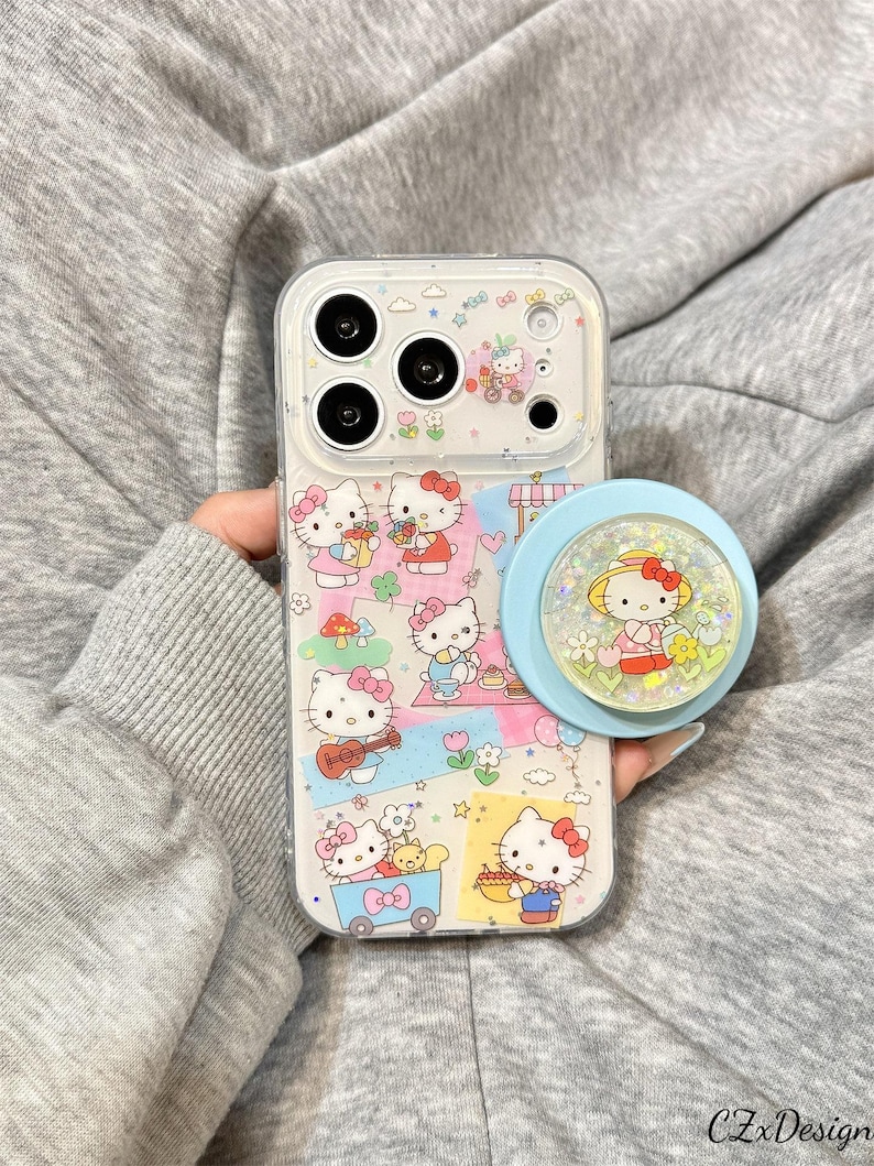 May include: Clear phone case with a colorful Hello Kitty design. The case features multiple illustrations of Hello Kitty in various scenes, along with a light blue pop socket with a Hello Kitty design. The case is held by a person.