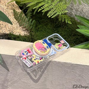 May include: Clear phone case with colorful cartoon character stickers and a pop-up grip. The case features a blue and pink design with a cartoon character on the grip. The case is decorated with various stickers.