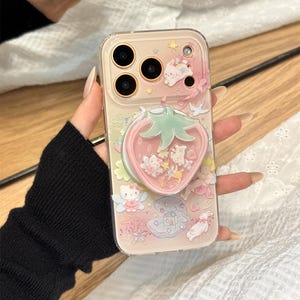 以下が含まれることがあります： イチゴ型のポップソケットが付いた透明な携帯電話ケース。ケースは、漫画のキャラクターと星が描かれたピンクと白のデザインです。ケースは透明な縁とピンクの背景を持っています。