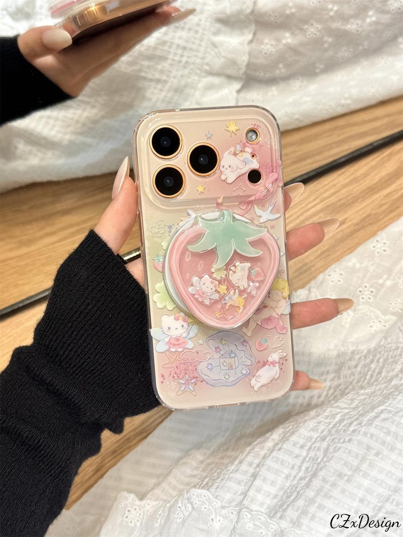 以下が含まれることがあります： イチゴ型のポップソケットが付いた透明な携帯電話ケース。ケースは、漫画のキャラクターと星が描かれたピンクと白のデザインです。ケースは透明な縁とピンクの背景を持っています。
