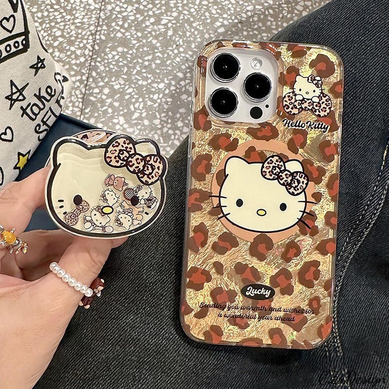May include: A clear phone case and a phone grip featuring a Hello Kitty design. The case has a gold leopard print background with a Hello Kitty graphic. The grip is shaped like Hello Kitty's face and has a bow.