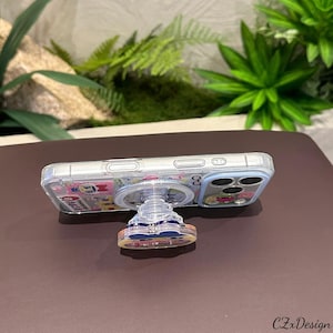 May include: A clear phone case with a pop-up grip featuring a colorful cartoon design. The phone case is on a brown surface with green plants in the background. The pop-up grip is round and has a clear base.