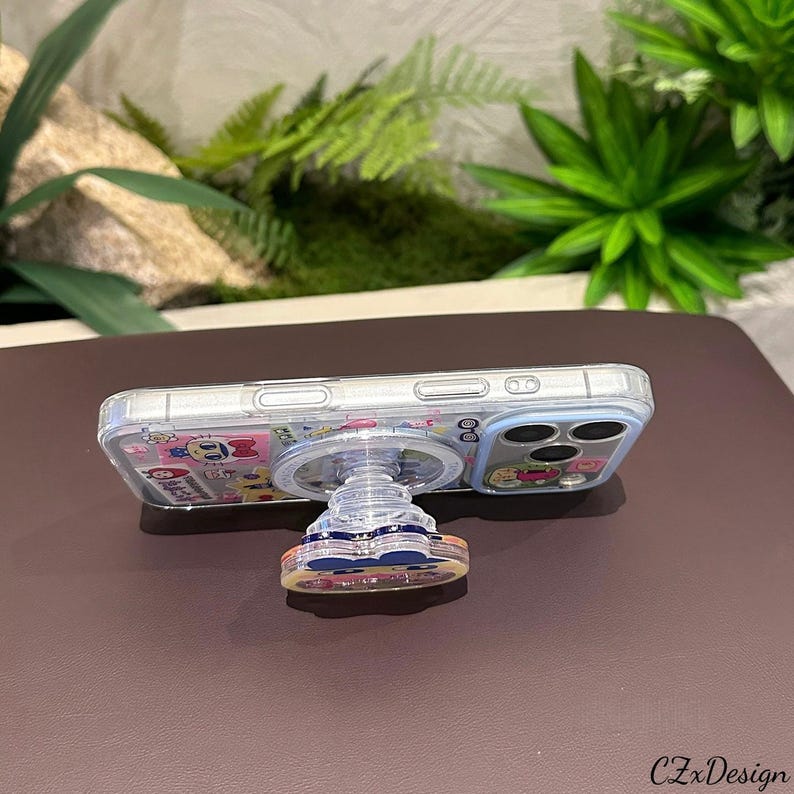 May include: A clear phone case with a pop-up grip featuring a colorful cartoon design. The phone case is on a brown surface with green plants in the background. The pop-up grip is round and has a clear base.