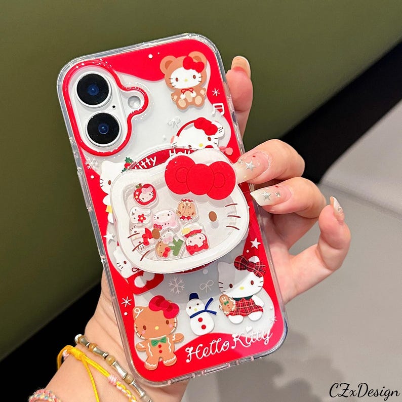May include: Red and clear phone case featuring Hello Kitty and other festive characters. The case has a pop-up grip with a red bow and various holiday-themed designs, including a gingerbread man and a snowman. The text "Hello Kitty" is visible.