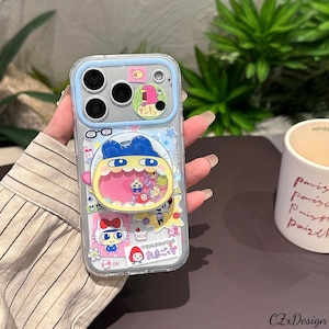 May include: Clear phone case with a blue border and a variety of colorful cartoon stickers. The stickers include a character with a large open mouth, a bird, and a Hello Kitty design. The case has a camera cutout and a pop-up grip.
