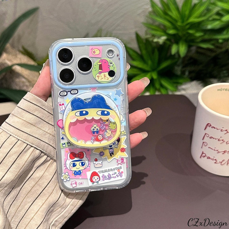 May include: Clear phone case with a blue border and a variety of colorful cartoon stickers. The stickers include a character with a large open mouth, a bird, and a Hello Kitty design. The case has a camera cutout and a pop-up grip.