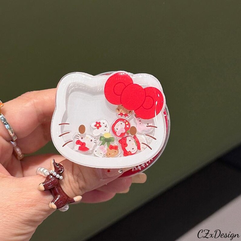 May include: A clear, round phone grip featuring a white Hello Kitty design with a red bow. The grip is filled with small, colorful floating charms. The design is set against a blurred green background.