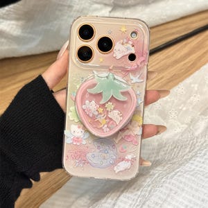 以下が含まれることがあります： ピンクとパステルカラーのデザインで、漫画のキャラクターとイチゴ型のポップソケットが特徴の透明な携帯電話ケース。ケースは、キラキラ光る液体入りのイチゴのデザインと透明なボーダーが特徴です。