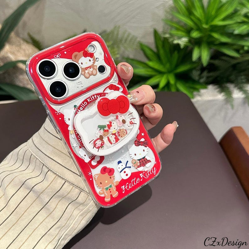 May include: Red and clear phone case featuring Hello Kitty characters. The case has a pop-up grip with a red bow design and the words "Hello Kitty". The case has a clear border and a red background with Hello Kitty graphics.