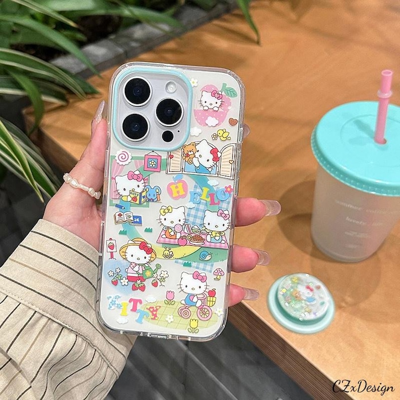 Pu&ograve; includere: Custodia trasparente per telefono con design Hello Kitty. La custodia ha un bordo azzurro ed &egrave; decorata con varie illustrazioni di Hello Kitty, tra cui scene di picnic e la parola "HELLO". Sono visibili anche un pop socket e una bevanda con cannuccia rosa.