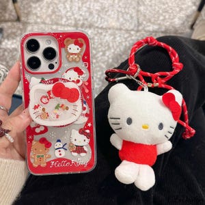 May include: A red and clear phone case with Hello Kitty designs and a matching plush keychain. The case features various Hello Kitty illustrations and the text "Hello Kitty". The plush keychain is white with a red bow and outfit.