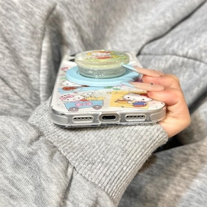 May include: Clear phone case with a cartoon design featuring a light blue pop socket. The case has a clear border and a colorful pattern with cartoon characters. The pop socket is light blue and clear.