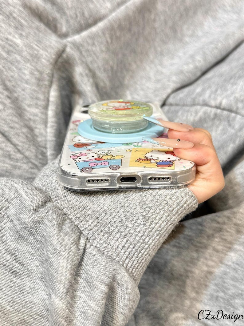 May include: Clear phone case with a cartoon design featuring a light blue pop socket. The case has a clear border and a colorful pattern with cartoon characters. The pop socket is light blue and clear.