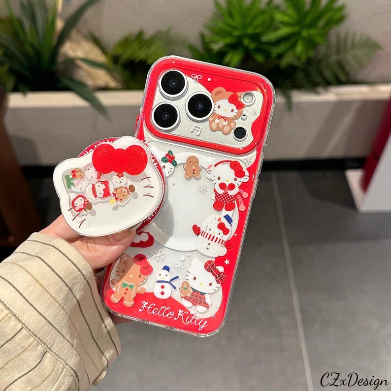 May include: Red and clear phone case with a Hello Kitty theme. The case features a red border and a white pop-up grip with a red bow. The case is decorated with Hello Kitty characters, gingerbread men, and snowflakes. The text "Hello Kitty" is visible.