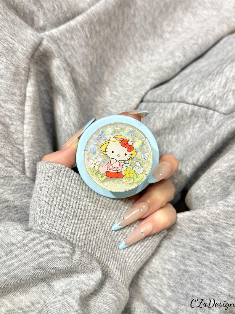 May include: A light blue compact mirror featuring a Hello Kitty design. The mirror has a clear, glitter-filled top and a cartoon image of Hello Kitty wearing a hat and dress. The mirror is held in a person's hand.
