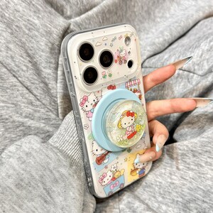 May include: Clear phone case featuring a colorful Hello Kitty design with a light blue pop socket. The case has a clear border and is decorated with various Hello Kitty illustrations and small details.
