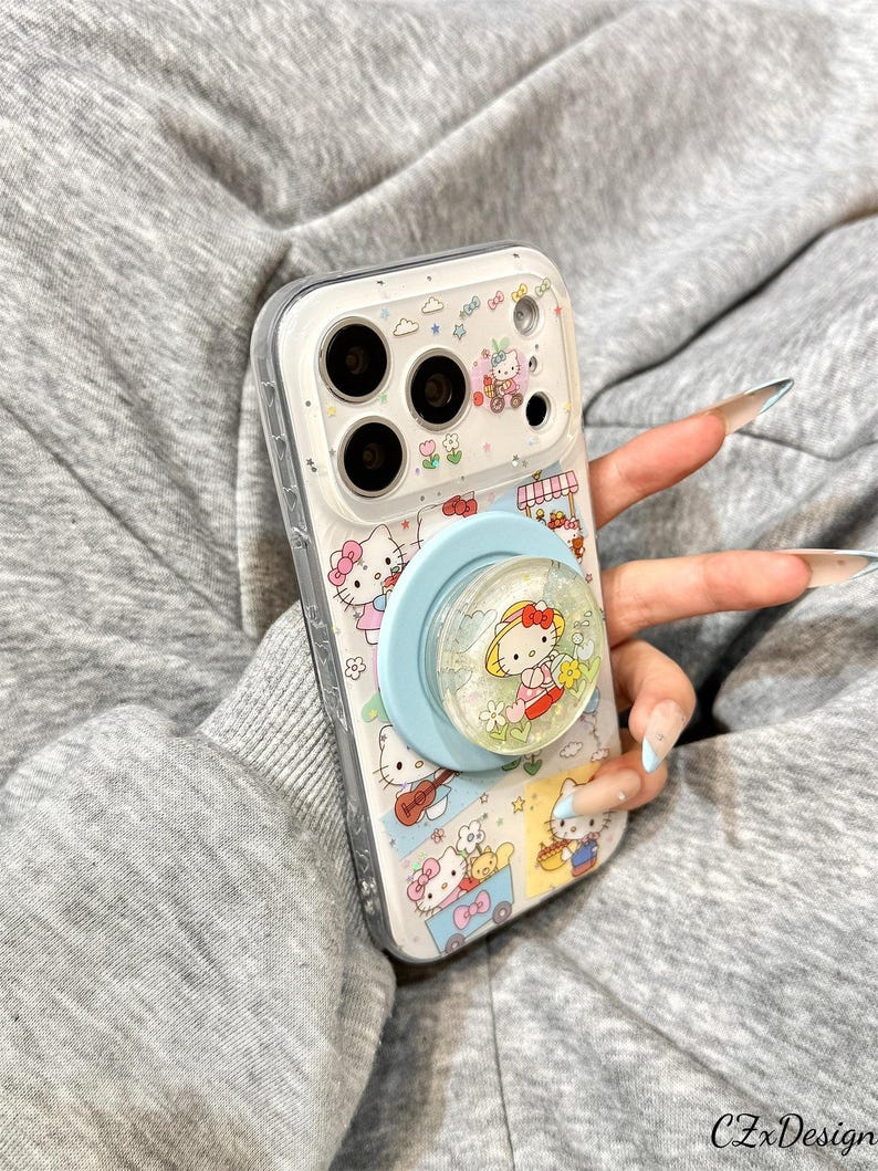 May include: Clear phone case featuring a colorful Hello Kitty design with a light blue pop socket. The case has a clear border and is decorated with various Hello Kitty illustrations and small details.