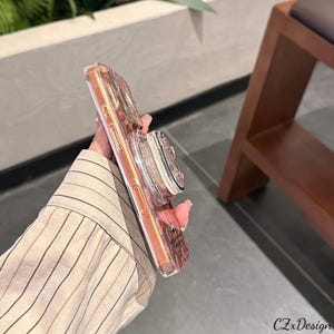 May include: A clear phone case with a rose gold border and a pop-up grip. The case has a design on the back and the pop-up grip is silver and white. The text "CZxDesign" is visible on the bottom right corner.