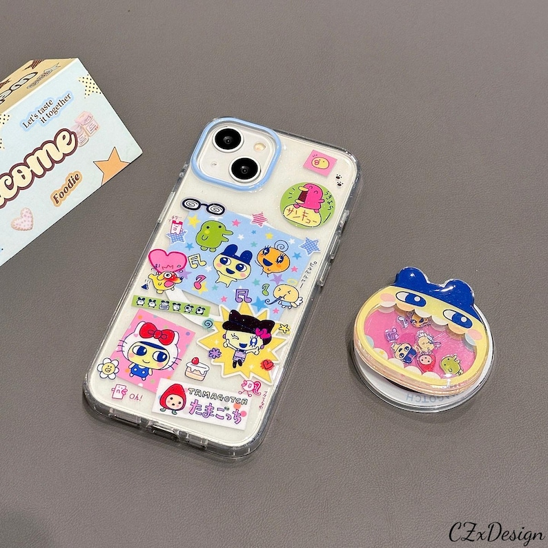 May include: Clear phone case decorated with colorful cartoon stickers. The stickers feature various characters and Japanese text. A matching phone grip with a cartoon character design is also present.