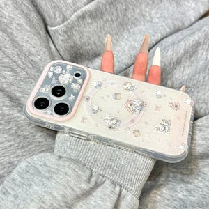 Może przedstawiać: Przezroczyste etui na telefon z r&oacute;żową obw&oacute;dką i kresk&oacute;wkowym wzorem przedstawiającym kr&oacute;liki, domki i inne małe ilustracje. Etui posiada wycięcie na aparat i pierścień MagSafe. Widoczny jest tekst "CZxDesign".
