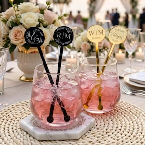Puede incluir: Dos vasos transparentes llenos de líquido rosa y cubitos de hielo, cada uno con dos agitadores de bebidas personalizados. Los agitadores negros muestran las iniciales A&M y R|M, mientras que los dorados muestran W|M 09.22.2023 y el nombre Villegas.