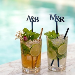 May include: Two cocktails in clear glasses, garnished with mint, lime slices, and decorative toppers with the letters "A&B" and "M&R". One drink has a gold straw and a pink rose. The other has a black straw.