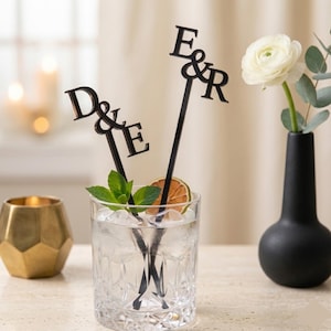 May include: Two black acrylic cocktail stirrers featuring custom lettering. One stirrer displays the letters D & E, while the other features the letters E & R. These decorative drink accessories are placed inside a clear glass filled with ice.