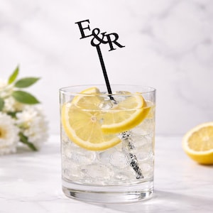 May include: A clear glass filled with ice and a lemon slice, with a black cocktail stirrer featuring the letters "E&R". A half lemon sits on the marble surface. The drink is a refreshing cocktail.