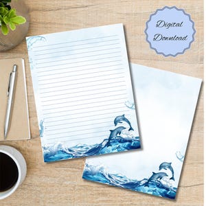 Puede incluir: Dos hojas de papelería con tema de delfines. El diseño presenta olas azules de acuarela y delfines saltando. Una hoja está rayada, la otra en blanco. Un sello azul claro dice "Descarga digital".