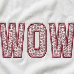 Op de afbeelding: Close-up van witte stof met het geborduurde woord "WOW". De letters zijn dieproze met een textuur, doolhofachtig patroon. Het lettertype is vet en schreef.