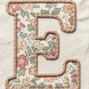 Puede incluir: Primer plano de una letra "E" de tela con un estampado floral. La letra está rellena de una tela color crema adornada con flores y hojas rosas, melocotón y verdes. Los bordes están delineados con un ribete trenzado rosa y verde.