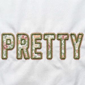May include: The word "PRETTY" embroidered on white fabric. The letters are filled with a floral pattern in pink, peach, and green, outlined with green stitching and a scalloped edge.