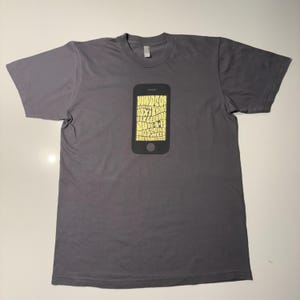 以下が含まれることがあります： 黒いスマートフォンと黄色のテキストが描かれたグレーのTシャツです。テキストには「WWDC08」、「OSX Leopard」、「Moscow」、「9-5」、「San Francisco」の文字が含まれています。このシャツは柔らかい素材で作られています。