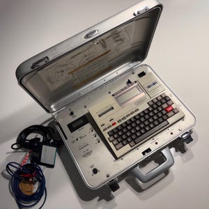Puede incluir: Un maletín plateado abierto que contiene una calculadora beige vintage con un teclado negro y teclas rojas. El maletín también contiene una pantalla digital, una ranura para cintas de cassette y un conjunto de cables y conectores.