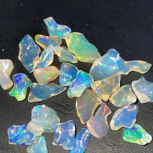 Könnte beinhalten: Eine Sammlung roher Opal-Edelsteine. Die Opale zeigen eine Reihe von Farben, darunter schillernde Blau-, Grün- und Gelbtöne, mit einer milchig-weißen Basis. Die Steine sind unregelmäßig geformt und variieren in der Größe, verstreut auf einer dunklen Oberfläche.