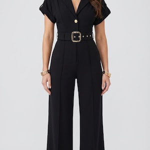 May include: A black jumpsuit with a collared neckline, short sleeves, and a V-neck. The jumpsuit features a gold buckle belt and a single gold button. The wide-leg pants are a solid black color. Gold bracelets adorn the wrists.