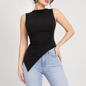 May include: Sleeveless black top with an asymmetrical hemline. The top is paired with light blue jeans. The model is wearing black sunglasses and has her hair up. The top has a ruched detail on the side.