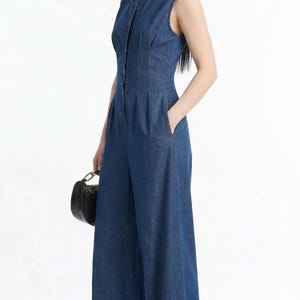 May include: Sleeveless denim jumpsuit in a medium blue wash. The jumpsuit features a collared neckline, button-down front, and wide-leg design. It includes front pockets and a fitted bodice. A small black handbag is held in the left hand.