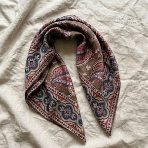 May include: A brown paisley scarf with red, blue, and pink patterns. The scarf is folded, displaying its detailed design. The text "Whispering Scarves" is visible in the upper right corner. The scarf is likely made of silk or a similar fabric.