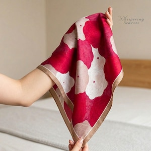 Peut inclure: Un carré de soie avec un motif floral. L'écharpe présente un fond rouge vif avec de grandes formes de fleurs blanches et rose clair. Les bords sont bordés d'une fine bordure beige. Le texte "Whispering Scarves" est visible dans le coin supérieur droit.