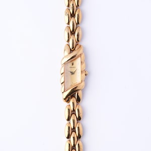 May include: A gold-toned Seiko wristwatch with a rectangular face and a linked bracelet. The watch face is a light gold color, and the band features an oval link design. The watch is set against a white background.