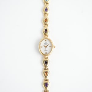 Puede incluir: Reloj de cuarzo dorado con esfera ovalada y blanca. La correa del reloj presenta piedras preciosas en forma de lágrima de varios colores. El reloj tiene números romanos y un pequeño segundero.