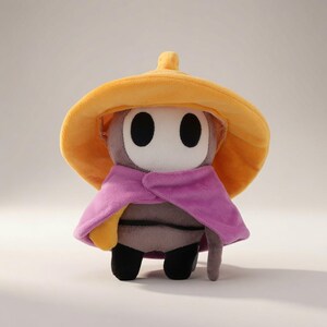 May include: A plush toy featuring a grey character with a white oval face and two black eye holes. The character wears a wide, soft yellow hat with a small point on top and a vibrant purple cape draped over its shoulders, covering its grey body.