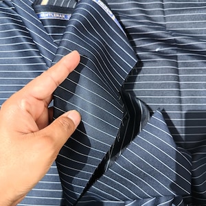 Può includere: Un tessuto blu navy scuro caratterizzato da sottili righe bianche uniformemente distanziate. Il materiale presenta un'etichetta visibile con il testo "GENTLEMAN" e "FIELIFE" stampato. Il tessuto è piegato, evidenziandone la trama liscia.