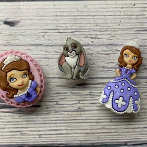 May include: Three princess-themed buttons. One button features a cartoon princess with a crown and purple dress. Another button features a cartoon rabbit with gray fur and pink ears. The third button features a cartoon princess with a crown and a purple dress with white polka dots.