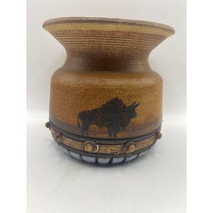 May include: A brown ceramic vase with a wide flared rim and a textured surface. The body features a black silhouette of a bison. A dark brown band with raised circular studs encircles the lower section, above a light blue base with vertical ridges.