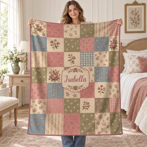 May include: A rectangular patchwork blanket featuring a grid of squares with various floral, striped, and checkered patterns in shades of pink, green, and blue. A central cream-colored oval plaque displays the name Isabella in script text.