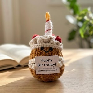 May include: A crocheted birthday cake with a candle and a small card that reads "Happy Birthday!" The cake is brown with white frosting and red berry accents. The candle has a yellow flame. The card also says "You deserve all the best today and always!"