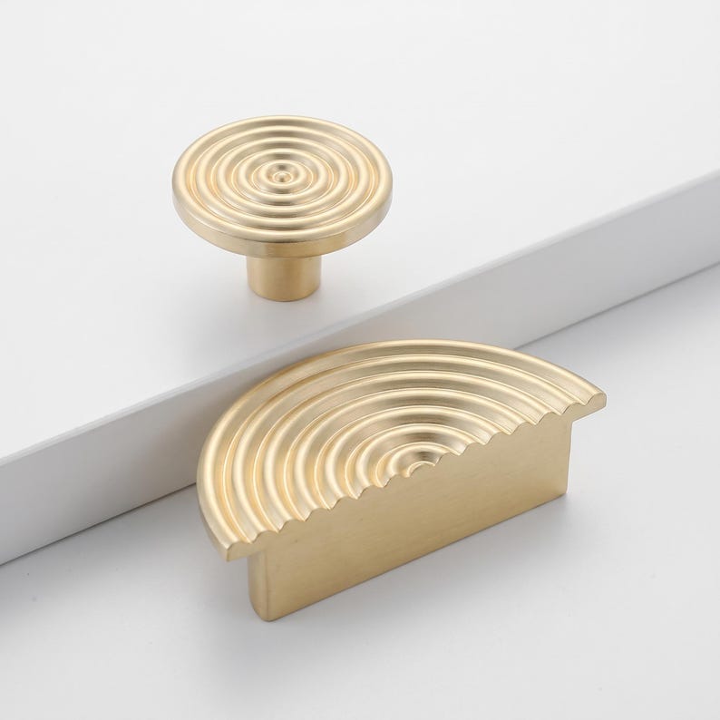 May include: A circular gold-toned cabinet knob and a semi-circular gold-toned cabinet pull, both featuring a concentric ripple pattern on their top surfaces. The circular knob has a cylindrical base, while the semi-circular pull has a flat edge.
