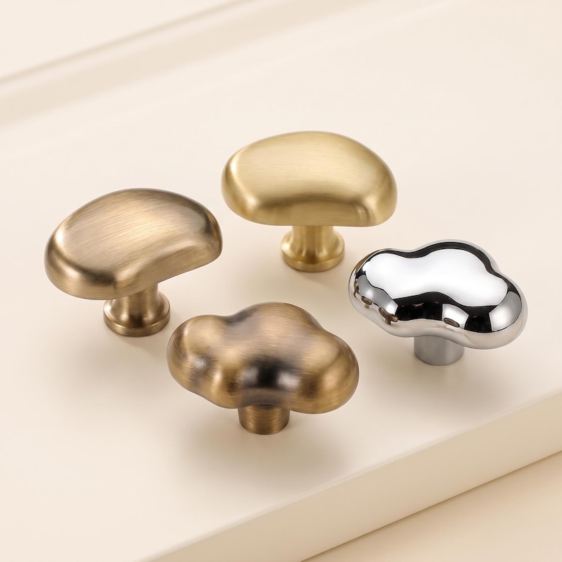 Op de afbeelding: Vier kastknoppen met onregelmatige, wolkachtige vormen. E&eacute;n knop heeft een gepolijste chroomafwerking, terwijl de andere drie vari&euml;rende metallic tinten van geborsteld messing en antiek brons vertonen. Elke knop heeft een centrale steel.